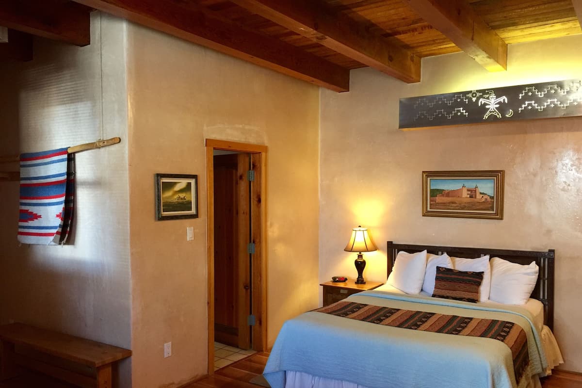 Quarto com cama de madeira, mesa de cabeceira com abajur, um banco e uma porta que dá para outro cômodo.