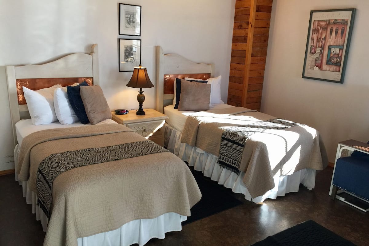 Quarto de hotel com duas camas, mesa de cabeceira com luminária, painel de parede em madeira e quadros na parede.