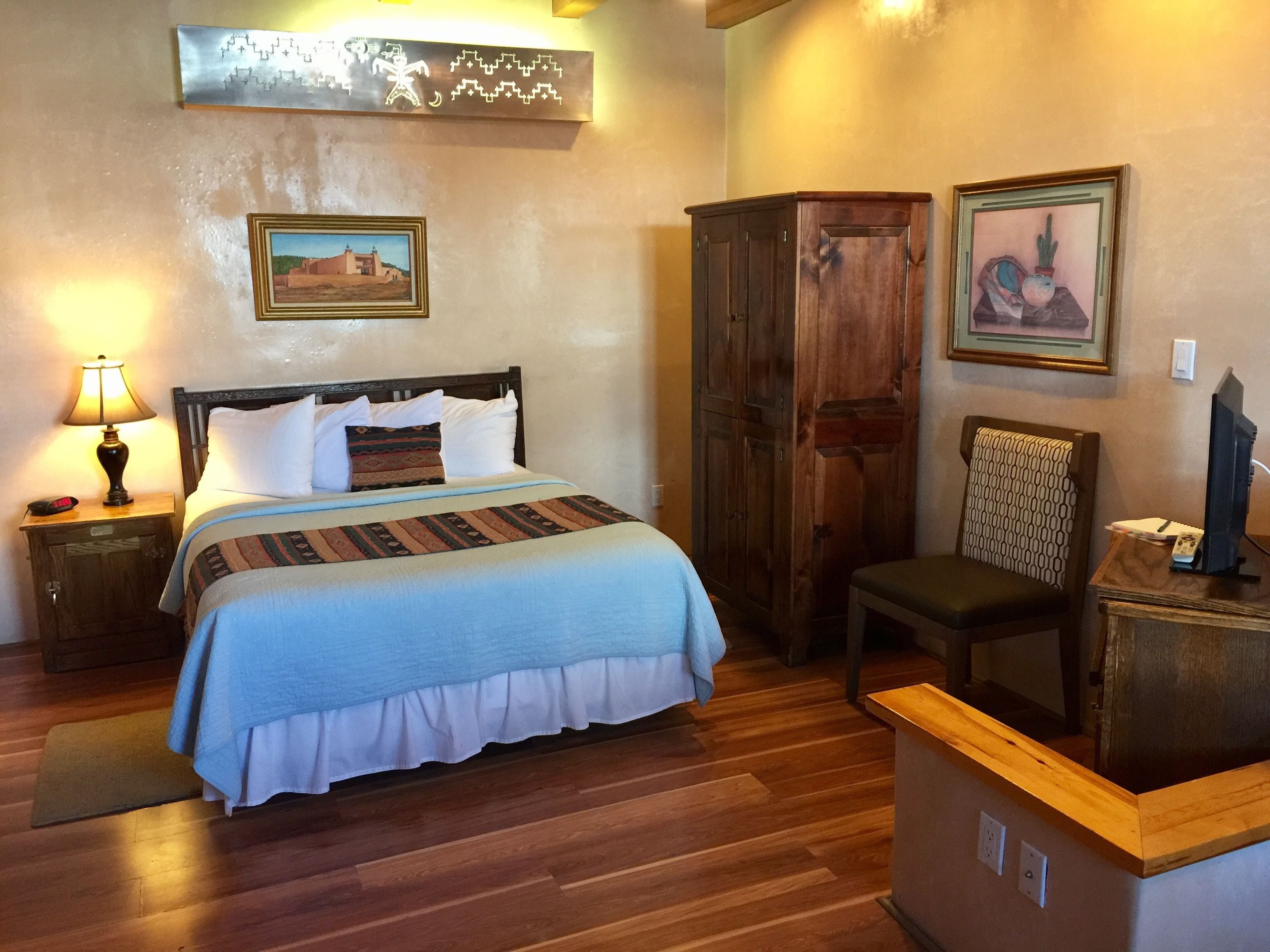 A bedroom with a bed, a nightstand, a wooden wardrobe, a chair, and a desk with a computer.