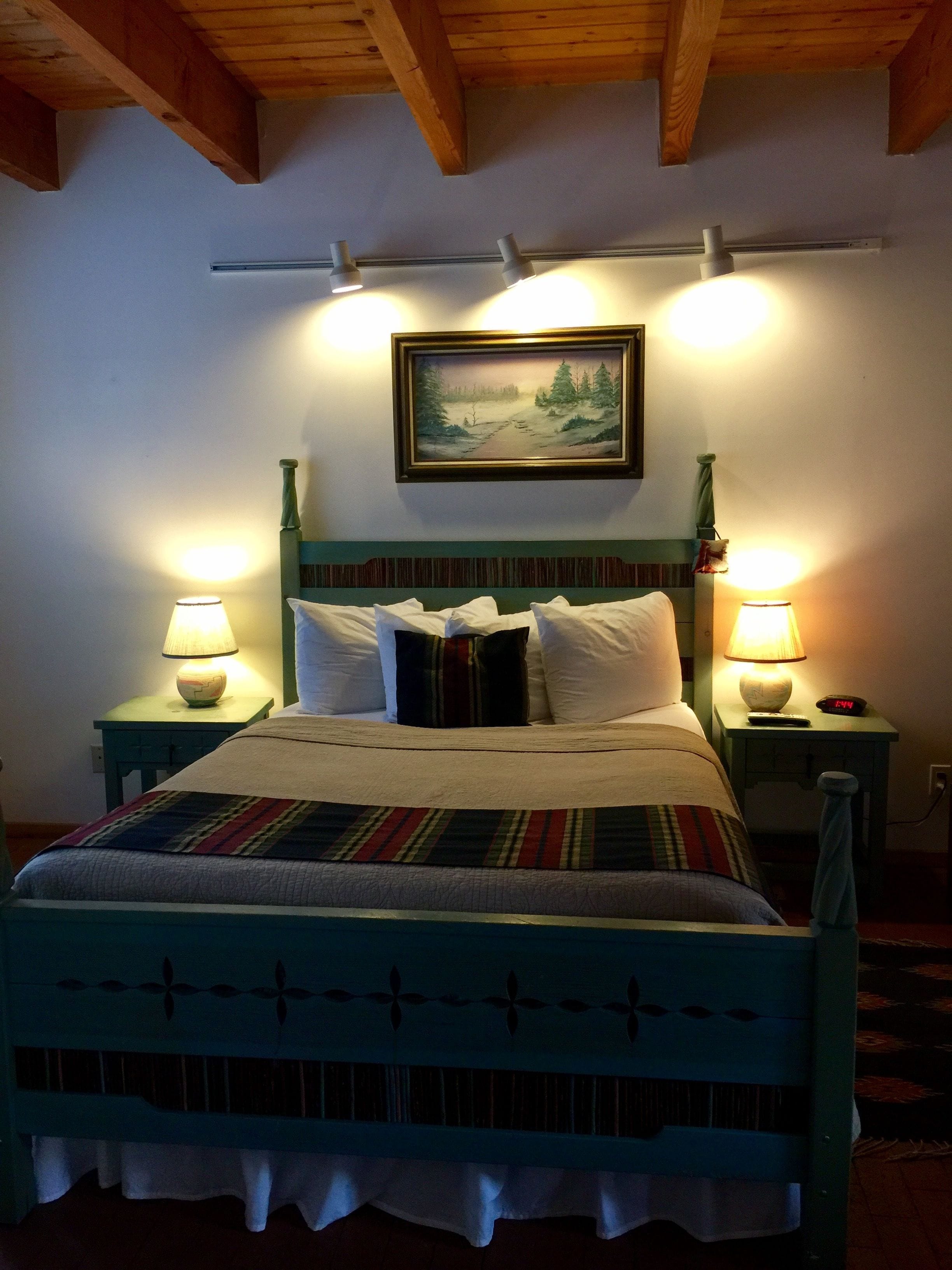 A bed with a striped blanket, two bedside lamps, a painting on the wall, and a wooden ceiling.