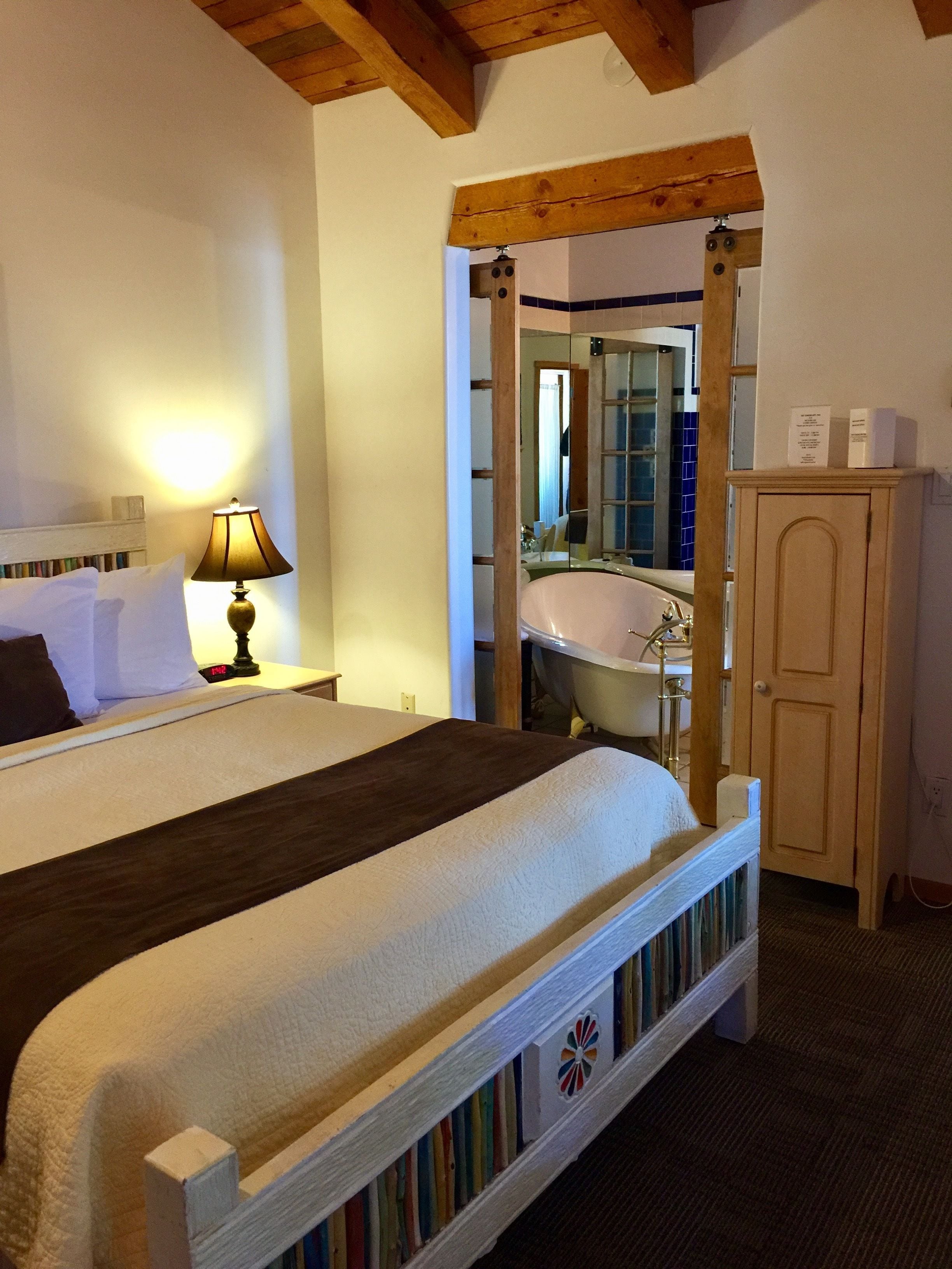 Una habitación de hotel con una cama, una mesita de noche con lámpara, un armario de madera y un baño visible a través de una puerta abierta.