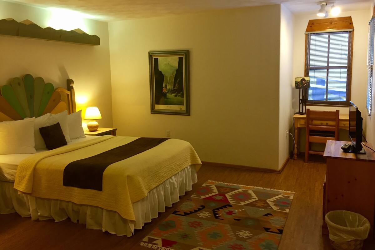 Quarto de hotel com cama, escrivaninha, cadeira, abajur, quadro na parede e janela com persianas.