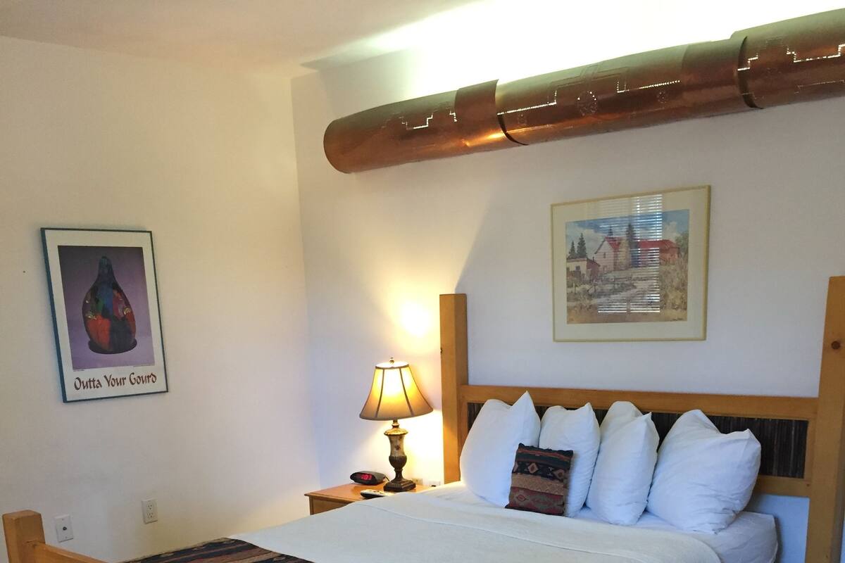Uma cama de madeira com colcha branca e várias almofadas, uma mesa de cabeceira com abajur e um quadro na parede.