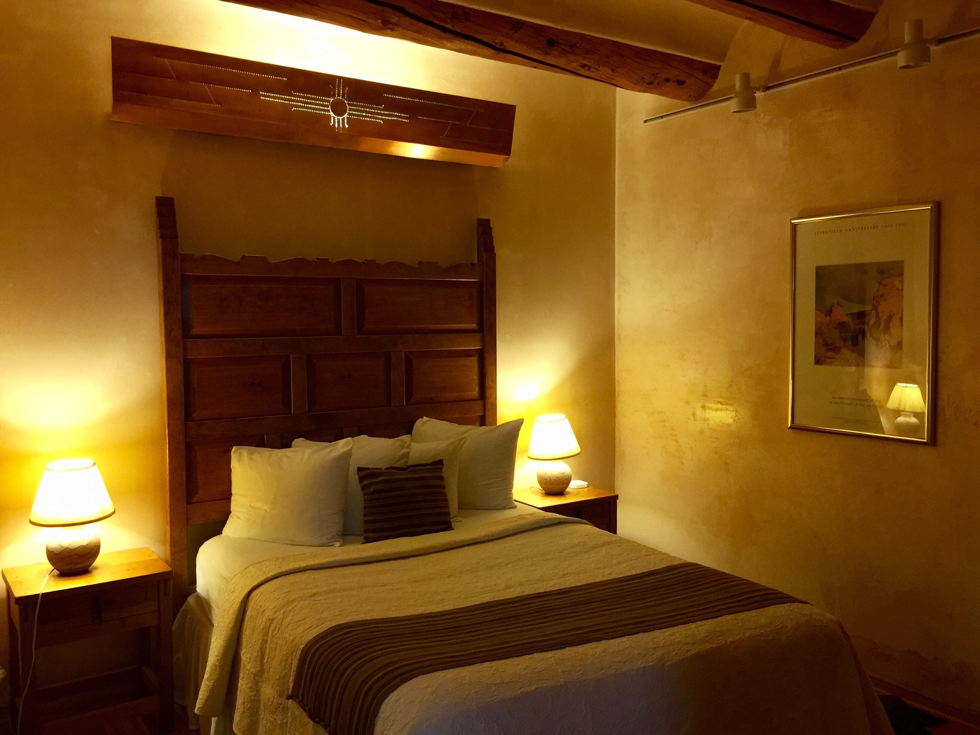 Habitación de hotel con cabecera de madera, dos lámparas de noche, un cuadro en la pared, y una cama con ropa blanca y una manta a rayas.