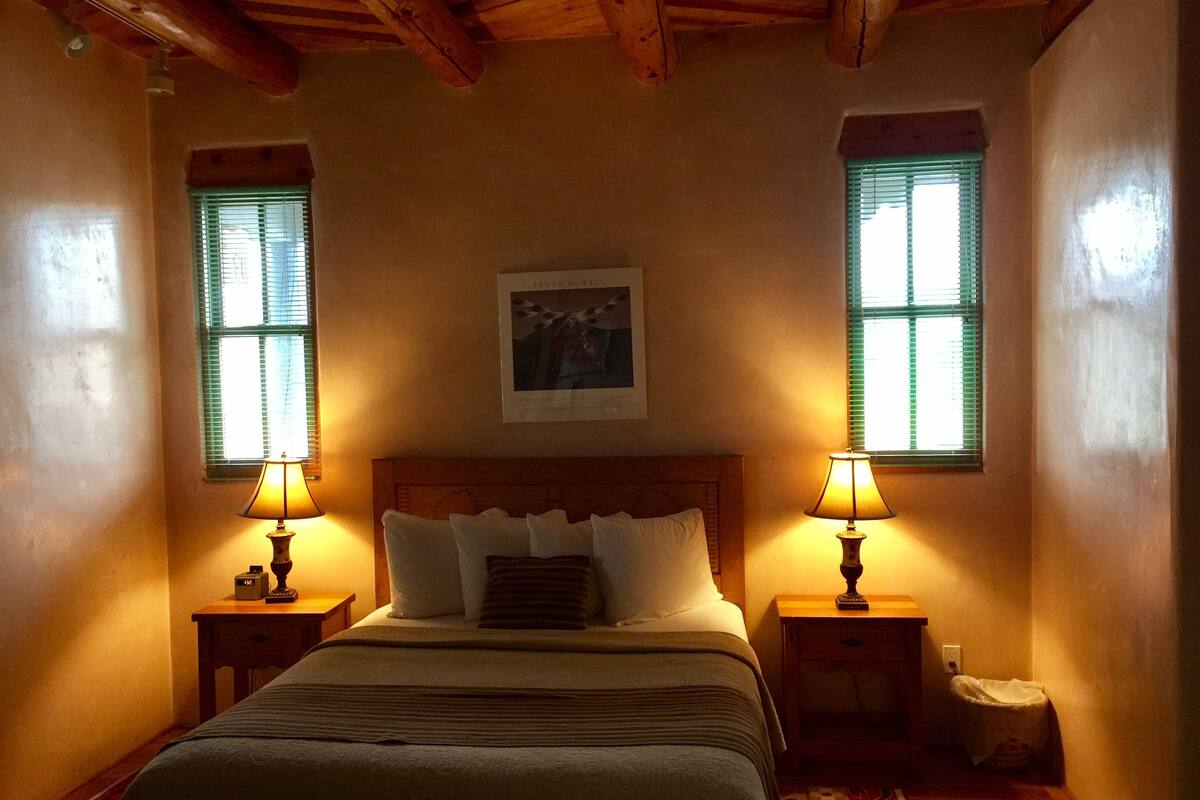 Quarto com cabeceira de madeira, duas luminárias de apoio, cama com várias almofadas e um quadro na parede.