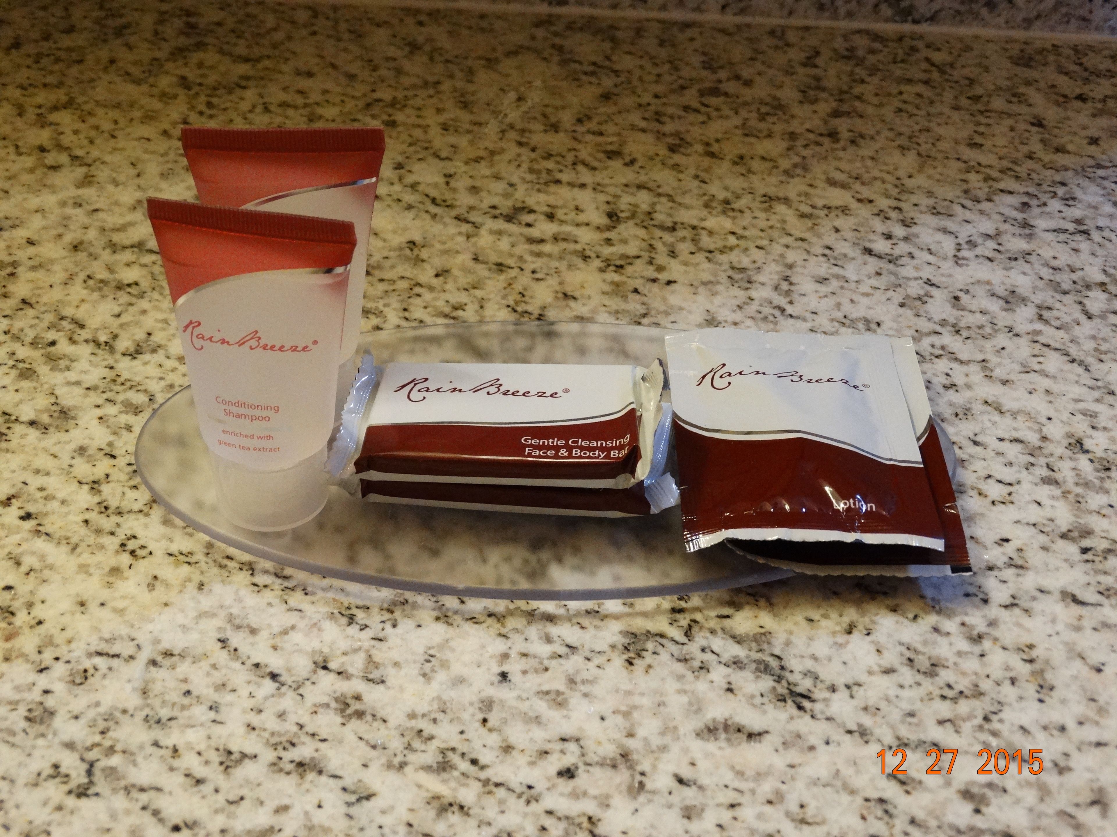 A countertop with a clear glass dish holding three items: a tube of RainBreeze Conditioning Shampoo, a bar of Gentle Cleansing Face & Body Bar, and a tube of RainBreeze Body Lotion.