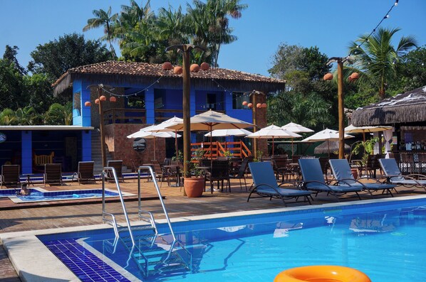 Zona de piscina con sillones de descanso, sombrillas y un edificio con tejado de baldosas.