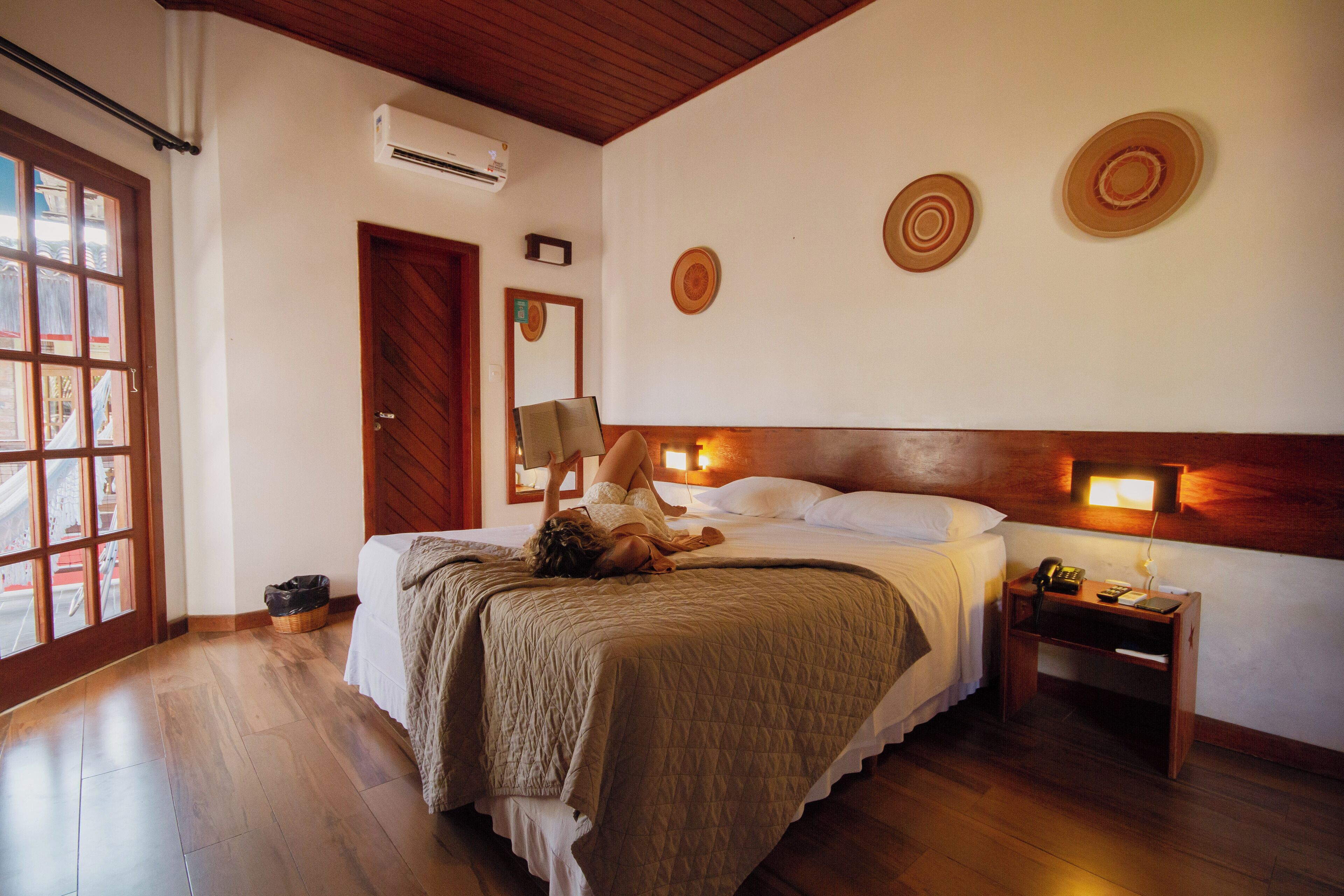 A hotel room with a wooden floor, a bed with a headboard, a bedside table with a lamp, and wall decor.