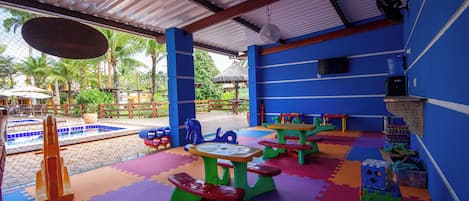 Un área de juegos interior con mesas y sillas coloridas, una piscina y una zona de estar cubierta.