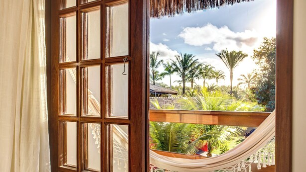 Vista desde una habitación con ventana de madera que da a una hamaca y abundante follaje.