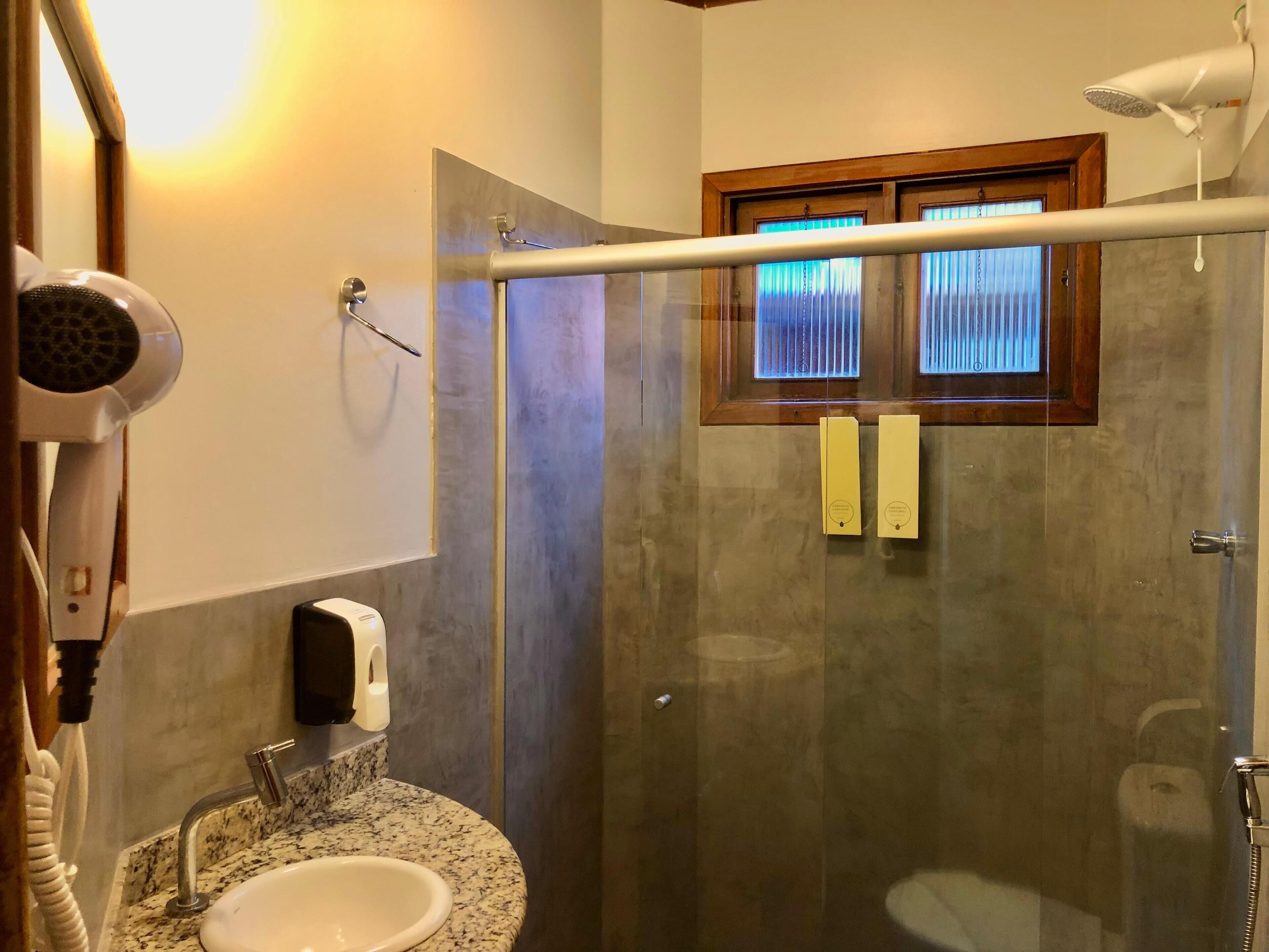A bathroom with a granite countertop sink, a handheld hair dryer, a towel dispenser, and a glass shower enclosure with a window.
