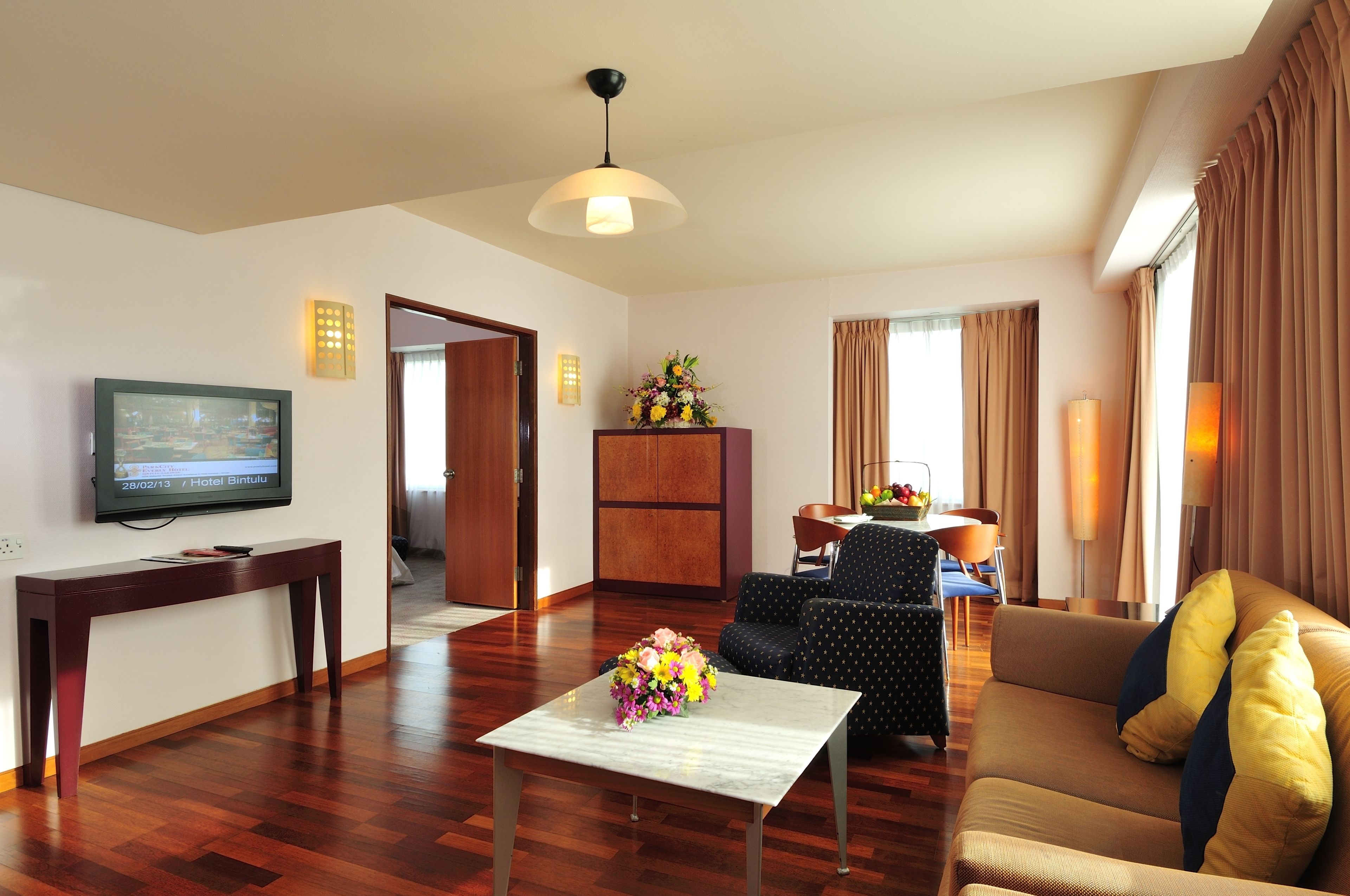A hotel room with a wooden floor, a TV on the wall, a sofa, a table with a vase of flowers, and a cabinet.