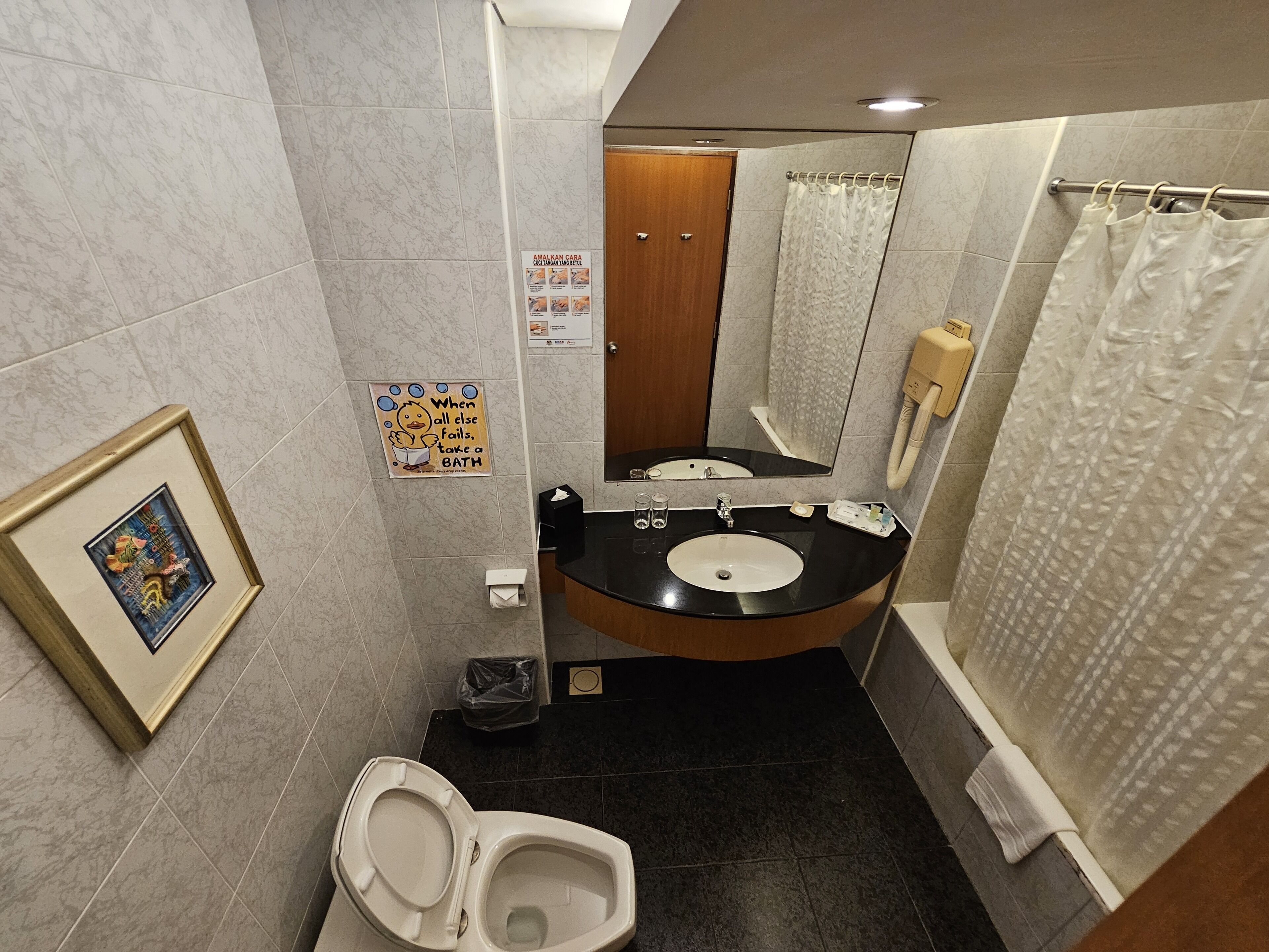 A bathroom with a toilet, sink, and shower area. There is a framed picture on the wall and a sign with a duck illustration.