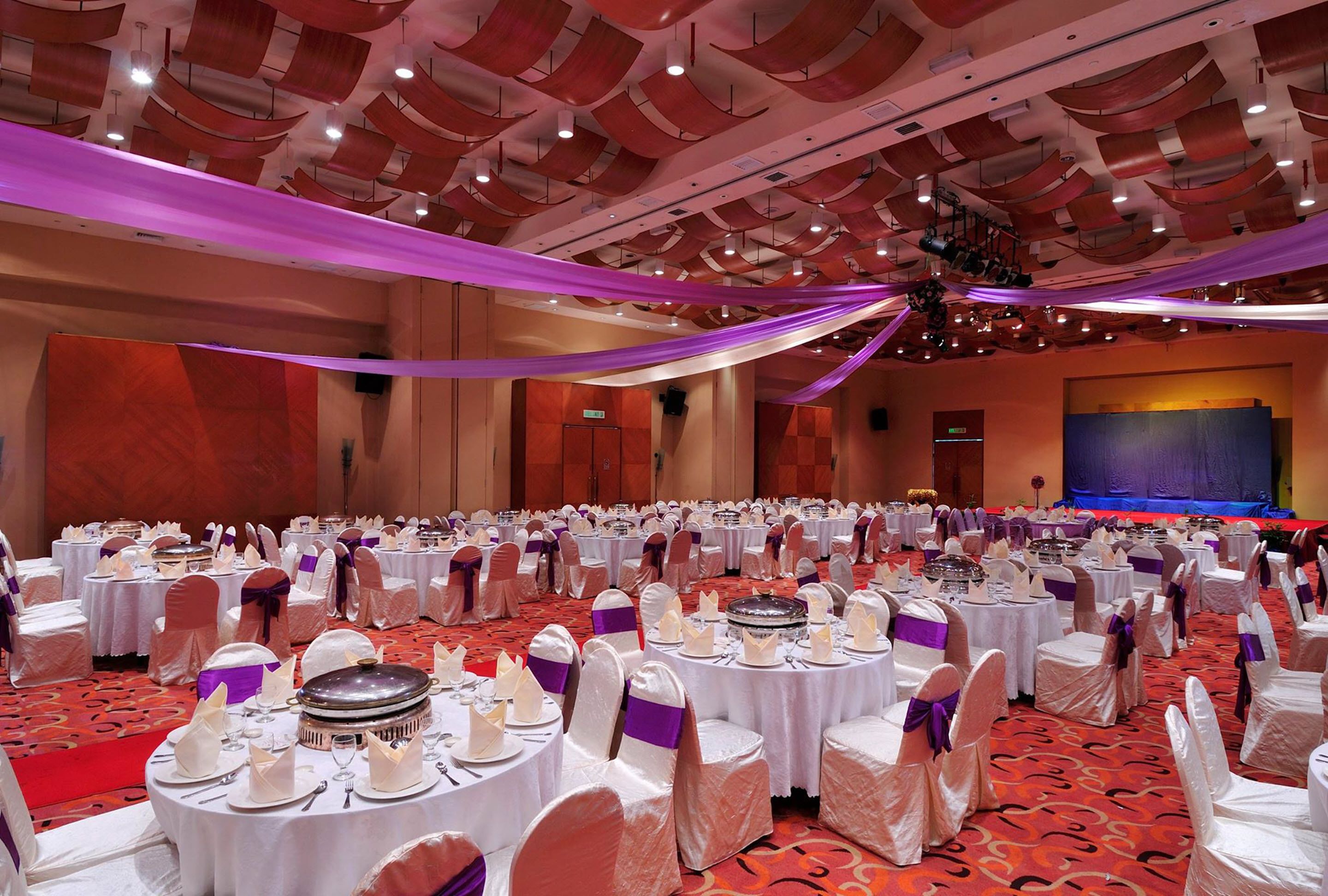 Un salón de banquetes con mesas redondas dispuestas para un evento formal, con manteles blancos y sillas adornadas con cintas moradas.