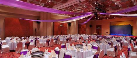 Un salón de banquetes con mesas redondas dispuestas para un evento formal, con manteles blancos y sillas adornadas con cintas moradas.
