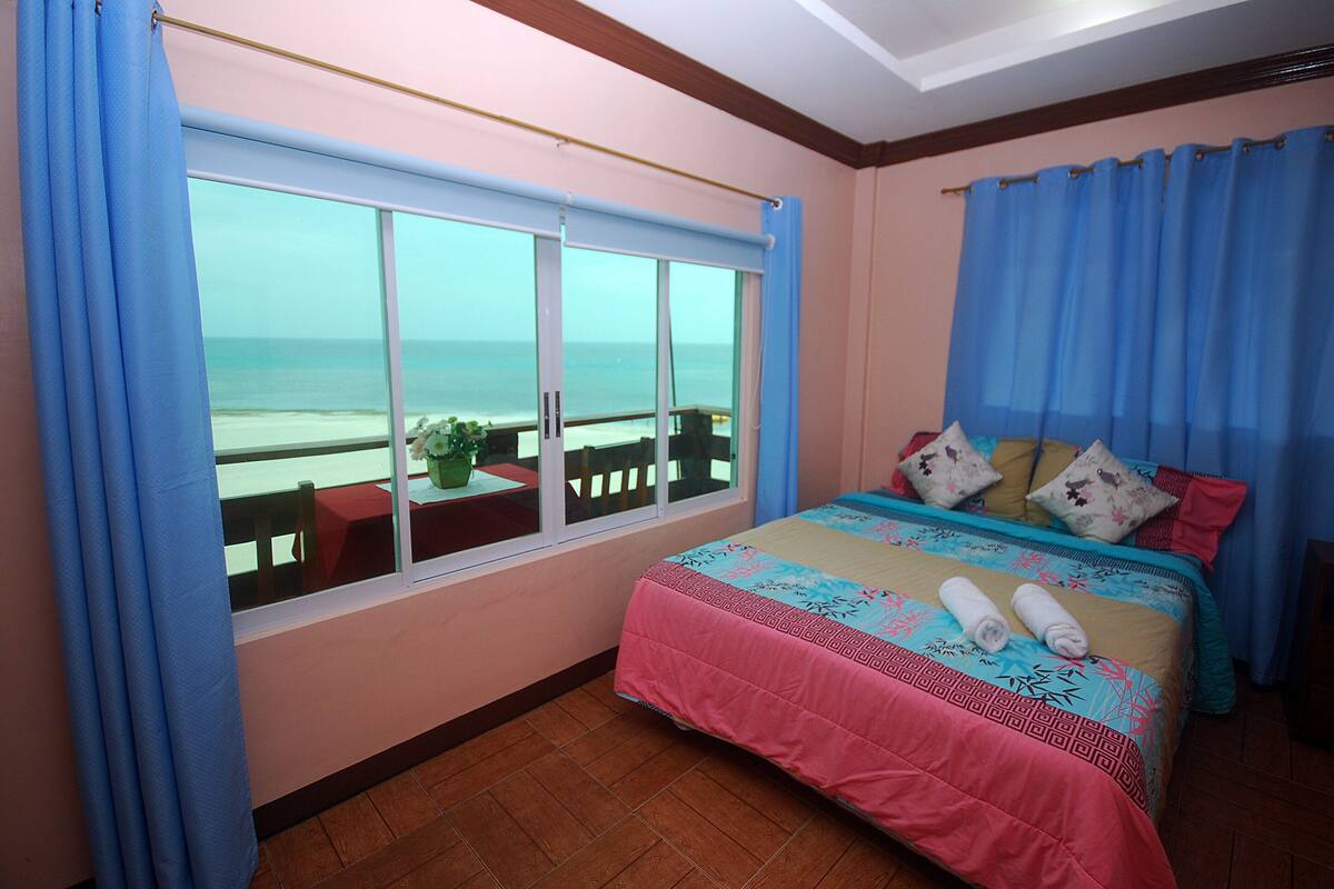 Ein Schlafzimmer mit großem Fenster mit Blick auf einen Strand, ein Bett mit Blumenmuster-Bettwäsche und Meerblick.