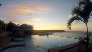 An infinity pool with a view of the sunset over the ocean, a palm tree, and a small building with a thatched roof. - Santiago Bay Garden & Resort (San Francisco)