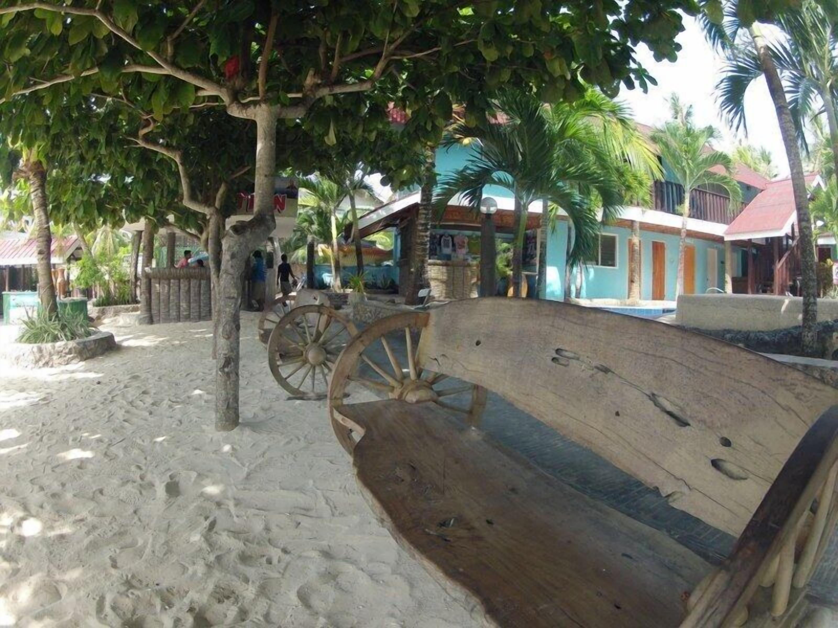 A sandy area with a wooden bench, palm trees, and a building with a red roof.