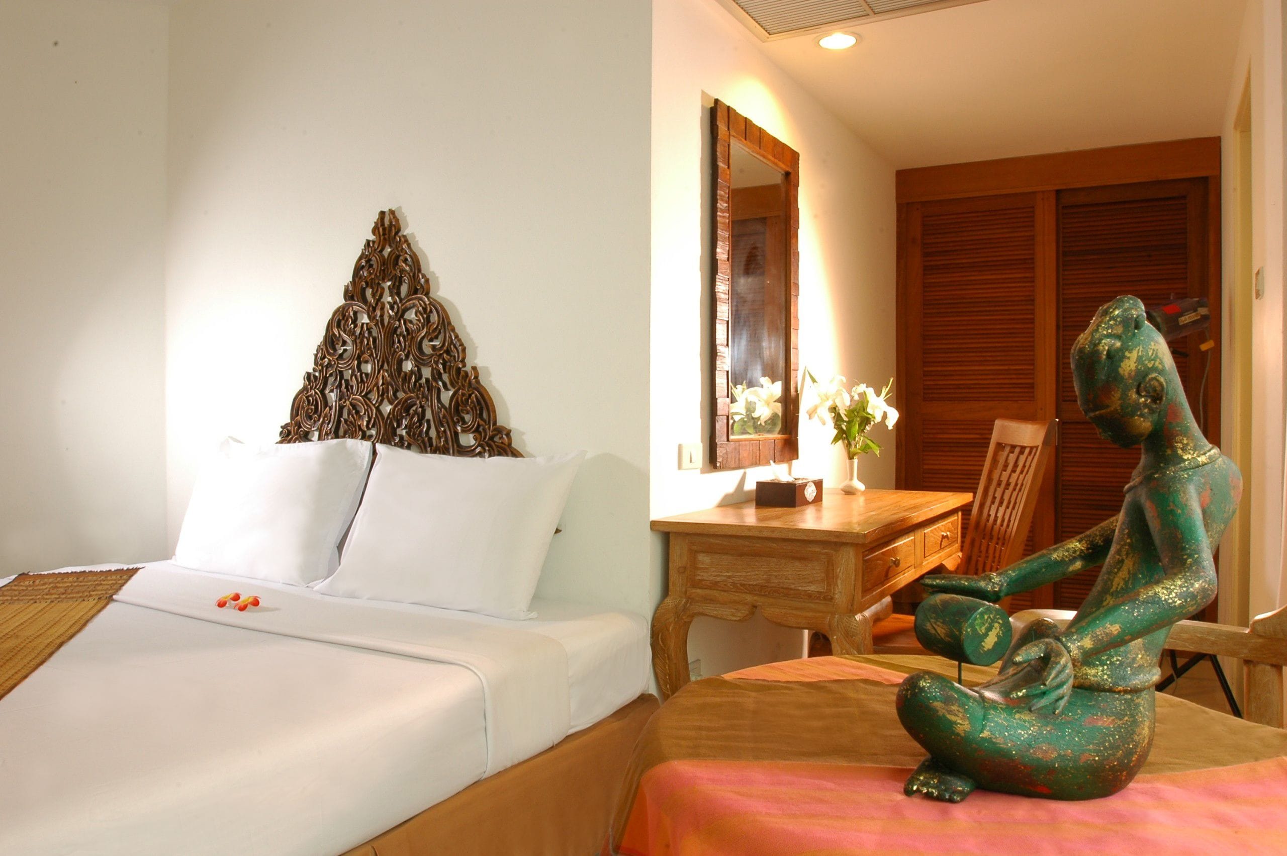 A hotel room with a bed, a wooden desk, a chair, a decorative wall piece, and a statue.