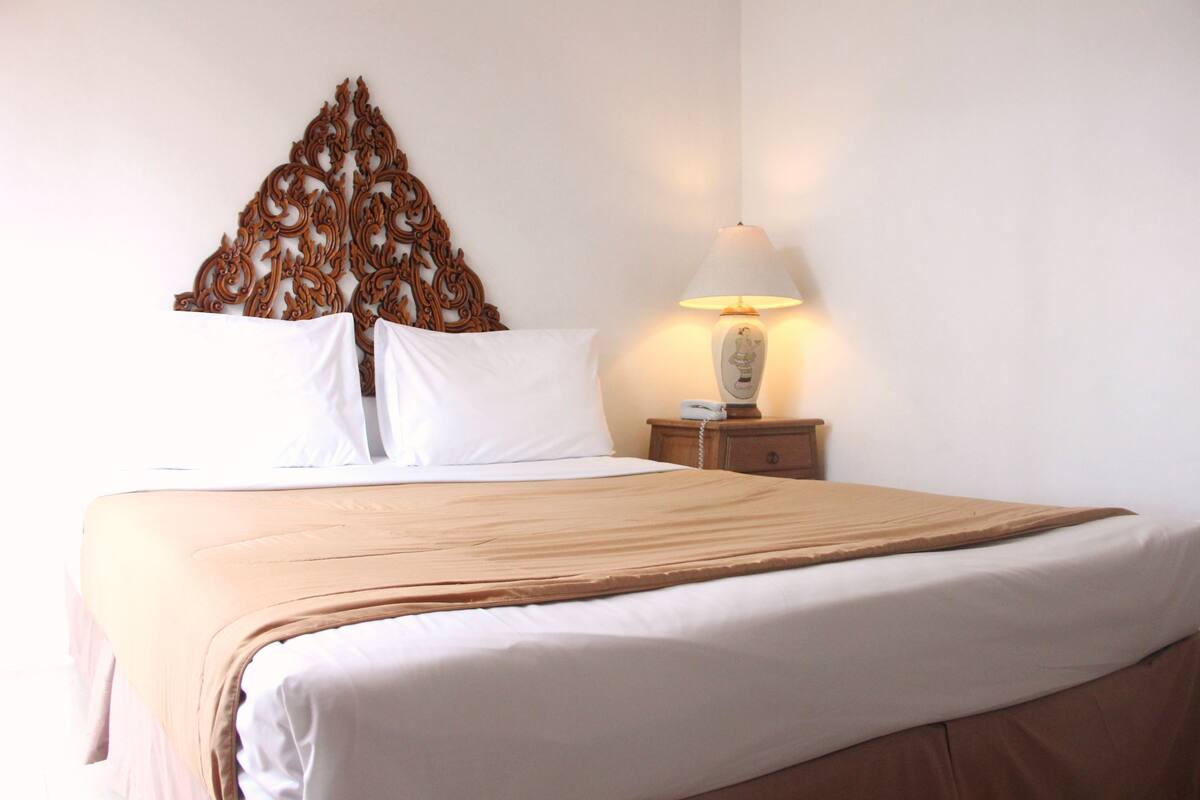 A neatly made bed with a beige bedspread and white pillows, a decorative wall piece above the bed, and a bedside table with a lamp.