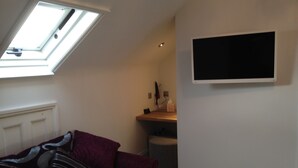 A room with a sloped ceiling, a sofa, a flat-screen TV mounted on the wall, and a small table with a lamp and a tissue box. - Brighton Inn Boutique Guest Accommodation (Brighton)