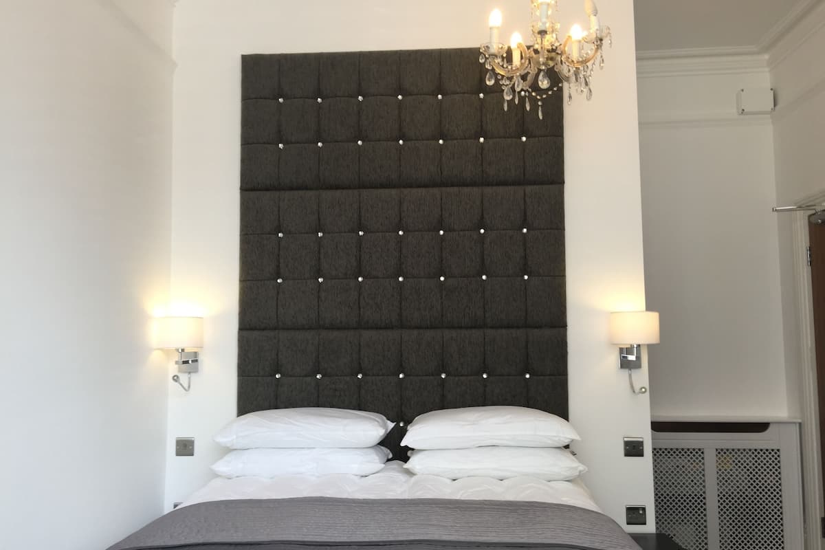 Un dormitorio con un cabecero capitonado, dos lámparas de noche, una araña y una cama con almohadas blancas y una colcha gris.