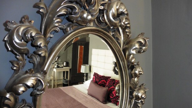 A room with a large ornate mirror reflecting a bed with a red and black patterned pillow, a bedside table with a lamp, and a chair. - Brighton Inn Boutique Guest Accommodation (Brighton)