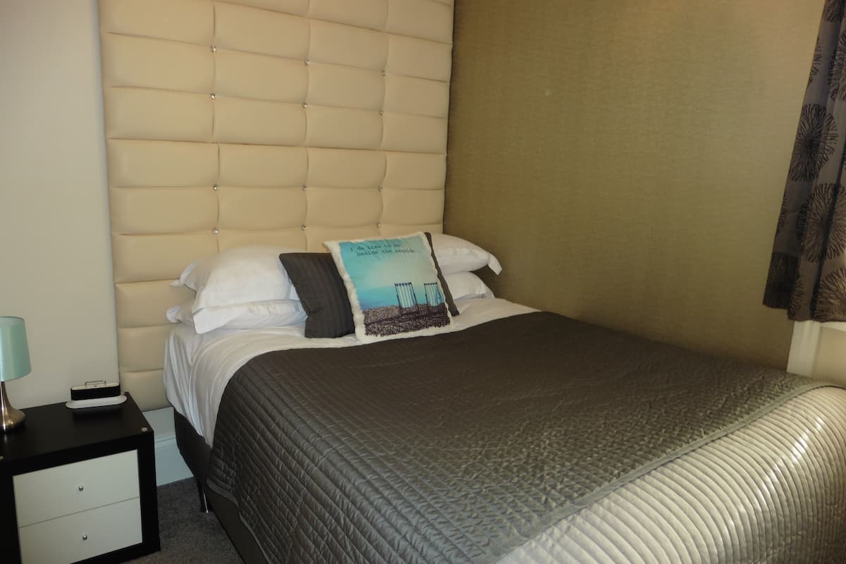 Una cama bien hecha con una colcha acolchada, un almohadón con estampado de paisaje y una mesita de noche con una lámpara y una pequeña caja.