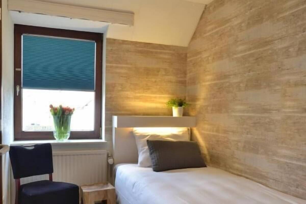 Ein Schlafzimmer mit Bett, Sessel, einem kleinen Tisch, einem Fenster mit Jalousien und einem an der Wand befestigten Regal mit Pflanze.