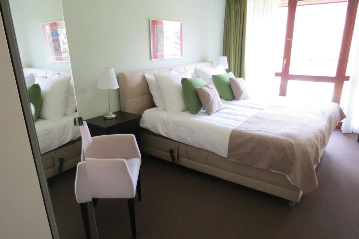 Ein Hotelzimmer mit zwei Betten, einem Sessel, einem Nachttisch und einem Fenster mit Vorhängen.