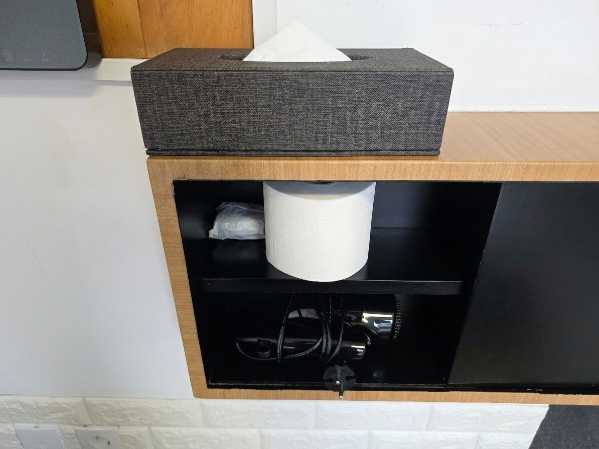 A black cabinet with a tissue box on top, a roll of toilet paper, and a black electrical cord.