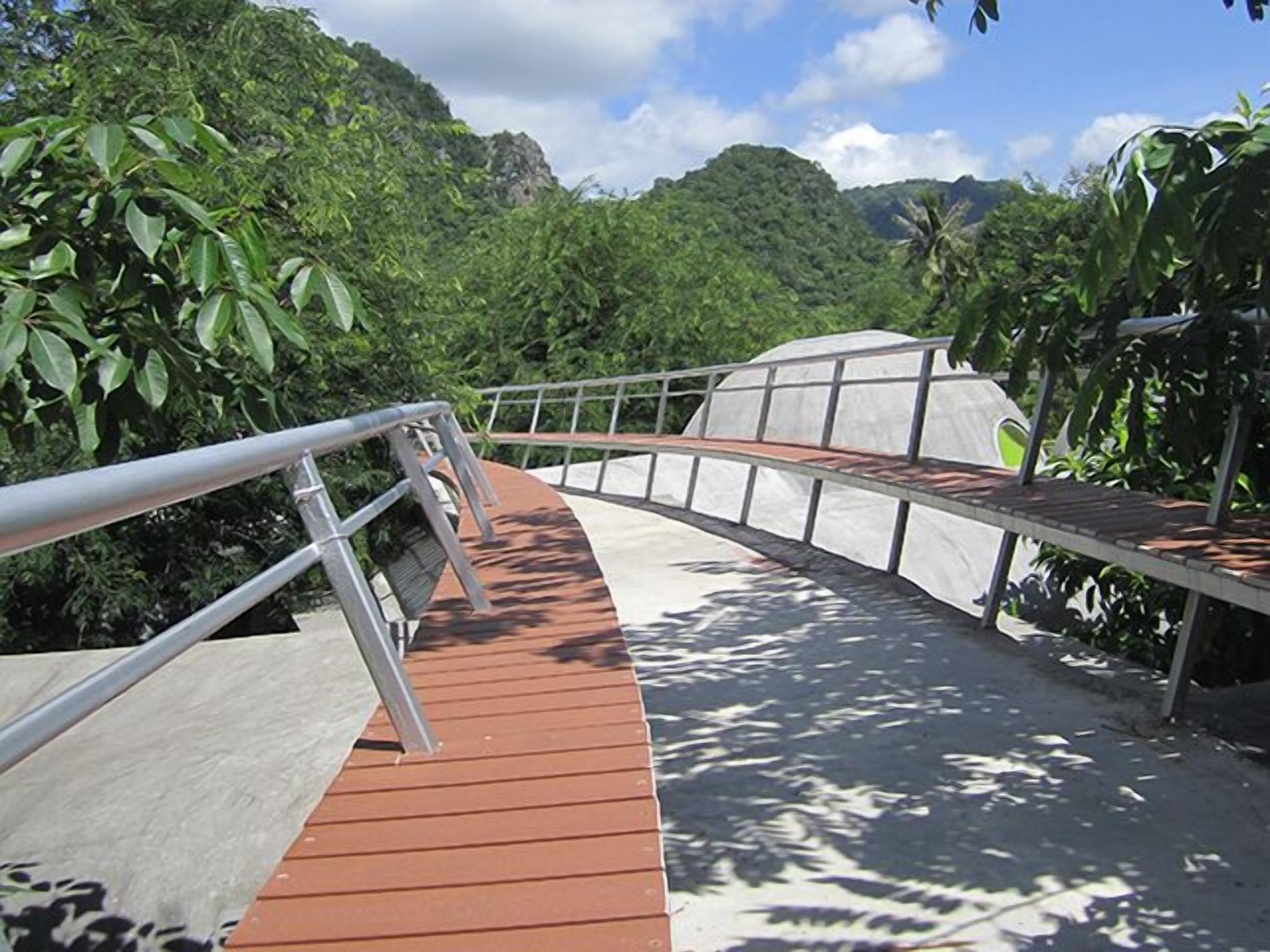 A wooden bridge with metal railings, surrounded by lush greenery and mountains.