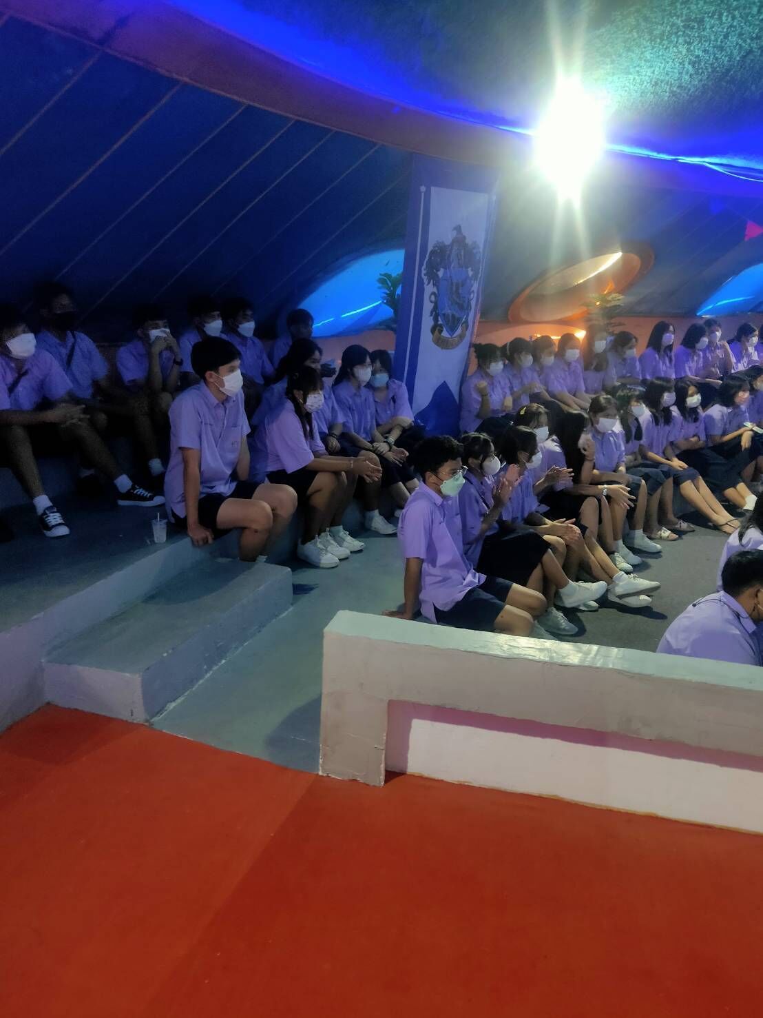 Un grupo de personas sentadas en un auditorio con una decoración azul y blanca, un banner con un escudo, y una fuente de luz brillante en el techo.