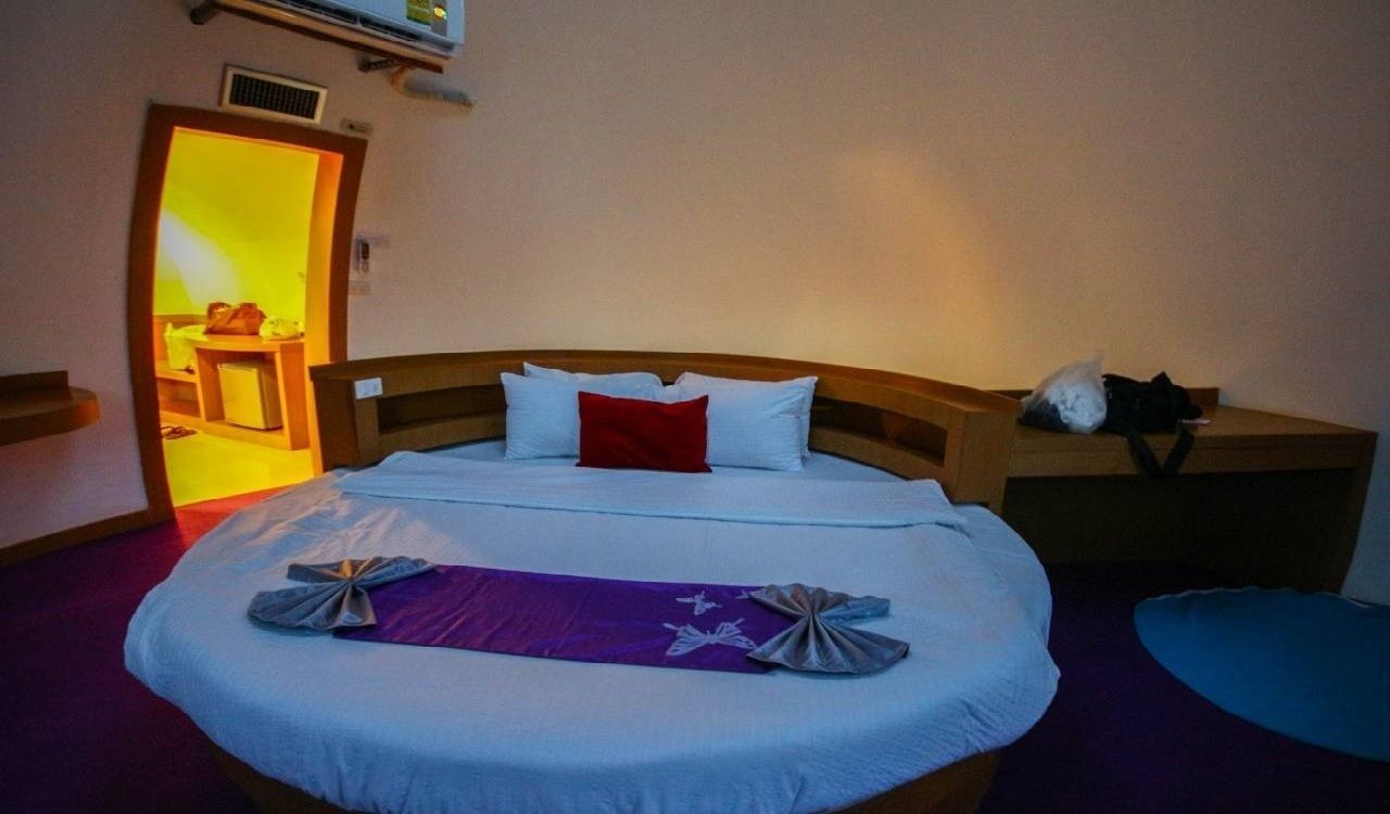 Un lit bien fait, avec une serviette violette et un oreiller blanc, une table de chevet en bois sur laquelle se trouve un sac noir, et une porte qui donne sur une pièce dont le mur est jaune.
