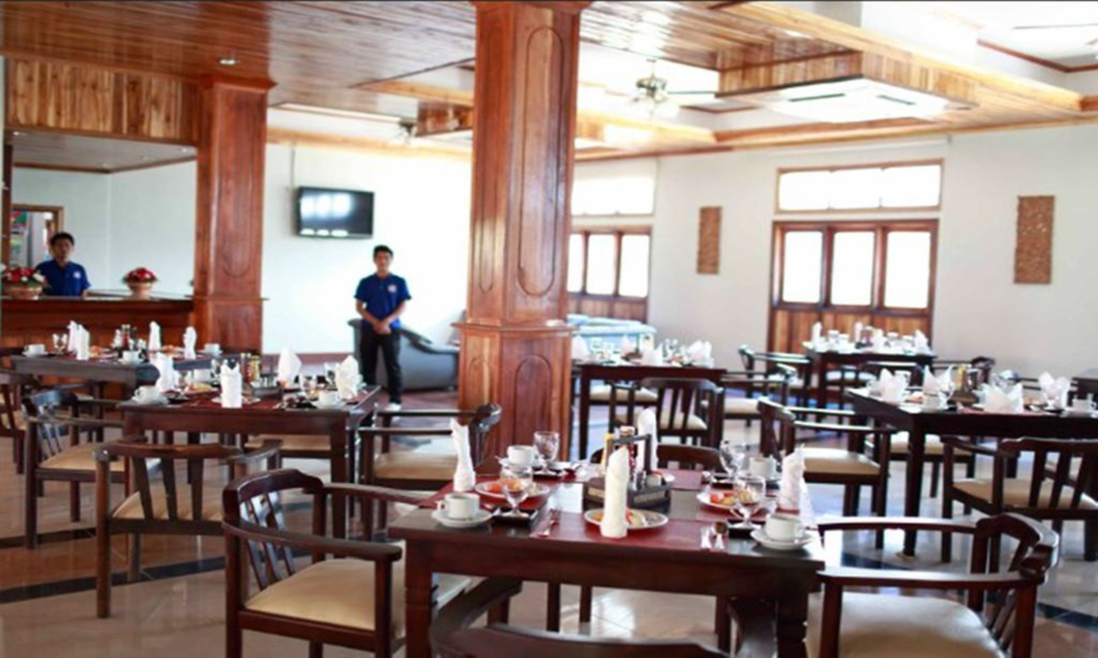 Un restaurante con mobiliario de madera y un techo alto. Las mesas están dispuestas con platos, vasos y cubiertos. Hay un televisor montado en la pared.