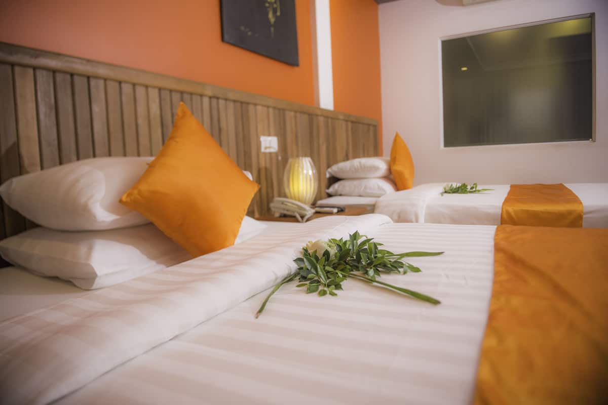 Habitación de hotel con dos camas, cada una con ropa de cama blanca y almohadas naranjas. Se observa un jarrón con flores sobre una cama, y una ventana que proporciona luz natural.
