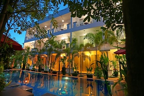 Um hotel com piscina, rodeado por vasos de plantas e guarda-sóis, com o edifício e a piscina refletidos na água.