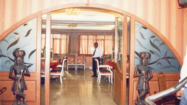 A room with a chandelier, a man standing, and a door with a sign above it. - Hotel TARA International (Hyderabad)