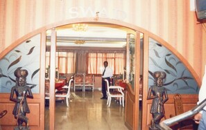 A room with a chandelier, a man standing, and a door with a sign above it. - Hotel TARA International (Hyderabad)