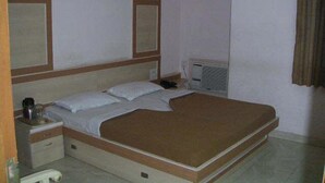 A standard single bed with a wooden headboard, a nightstand, and a bedside table. There is a window with a view of the outside, and a wall-mounted air conditioning unit. - Hotel TARA International (Hyderabad)