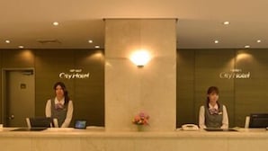 Un mostrador de recepción con dos recepcionistas, una placa con el nombre y un jarrón con flores.