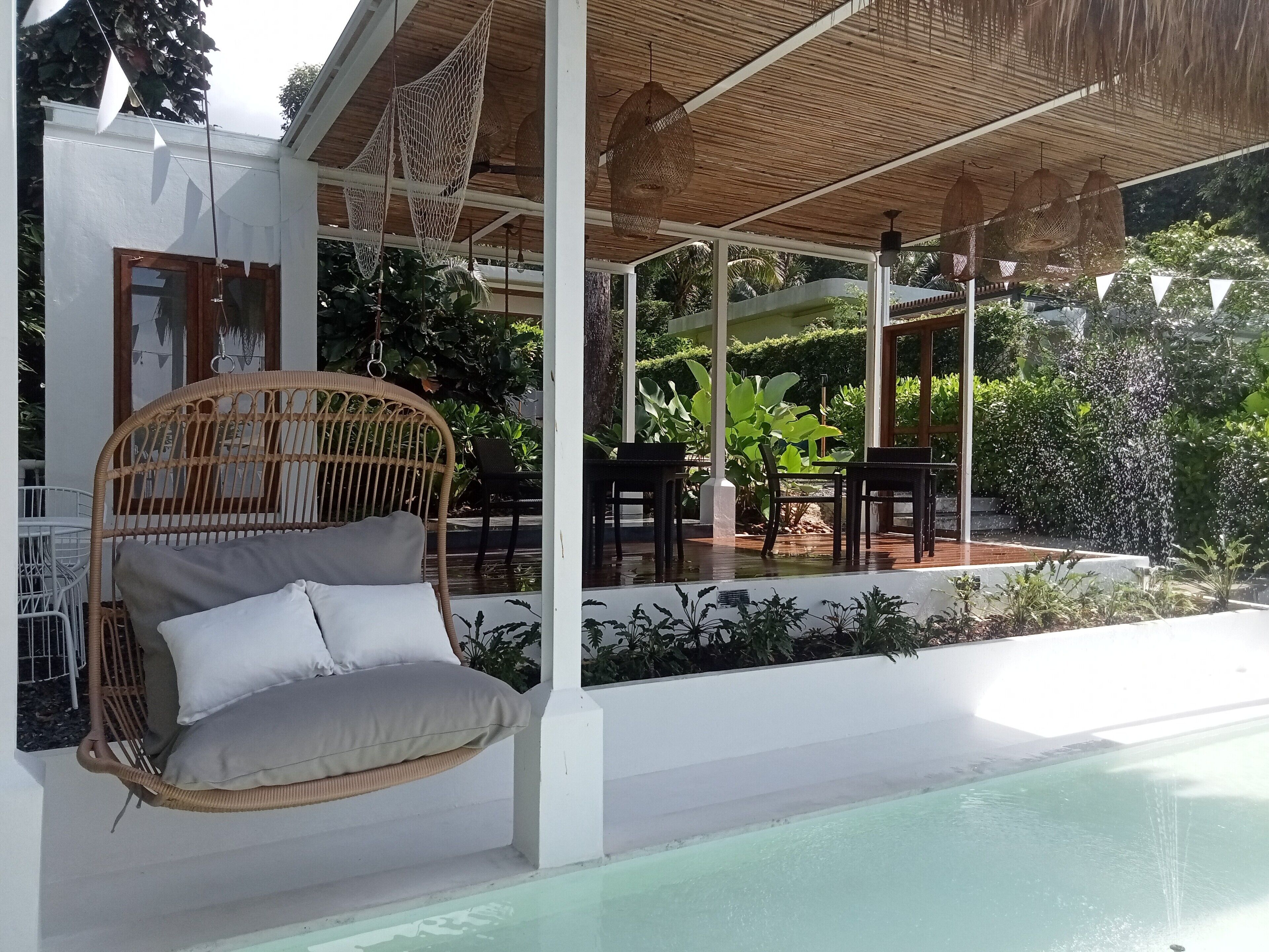 Un espacio de estar al aire libre con techo, que incluye una silla de mimbre con cojines, una piscina y abundante vegetación.