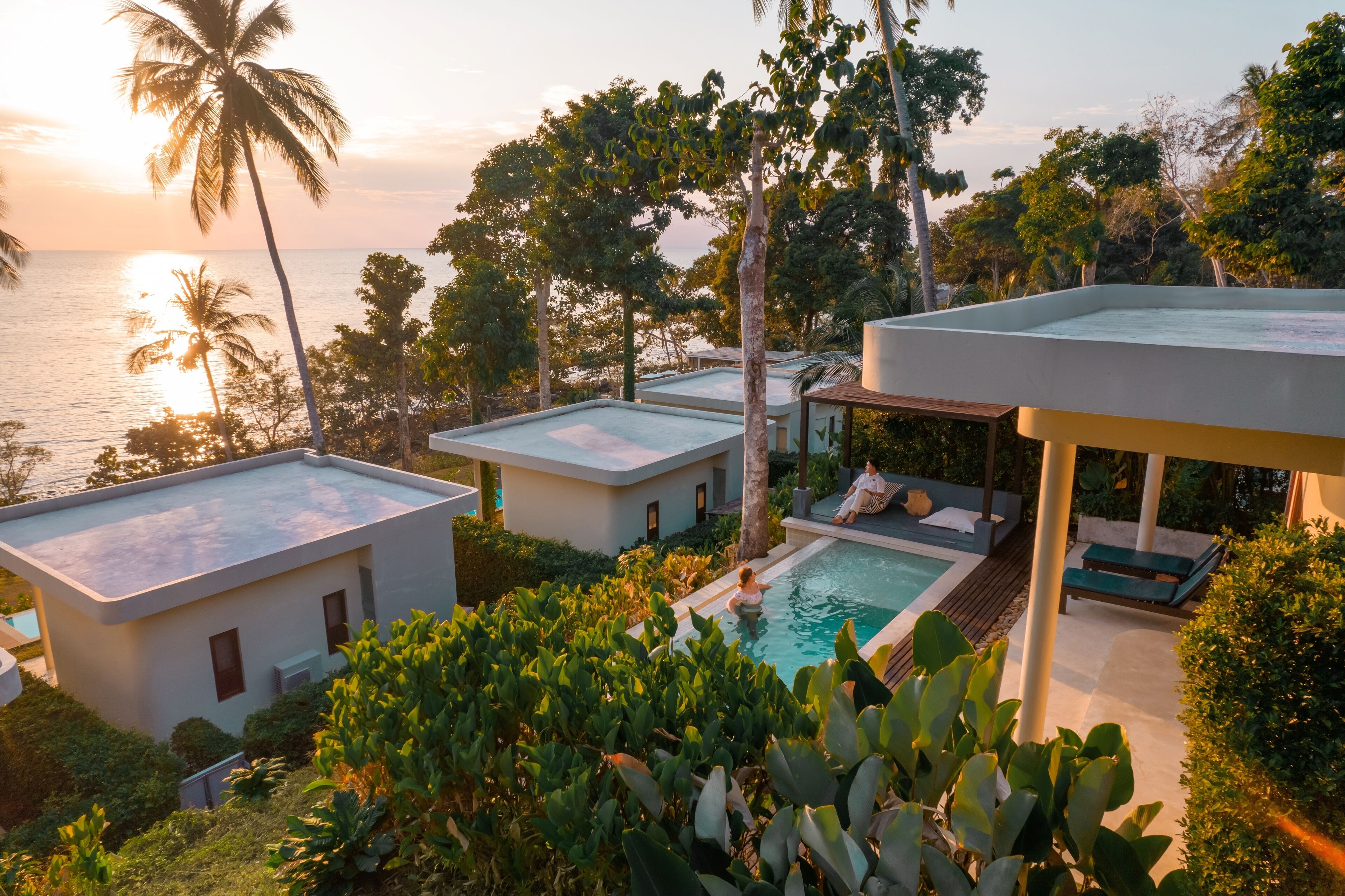 A modern resort with a pool, palm trees, and a view of the ocean at sunset.