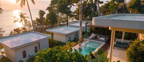 A modern resort with a pool, palm trees, and a view of the ocean at sunset.