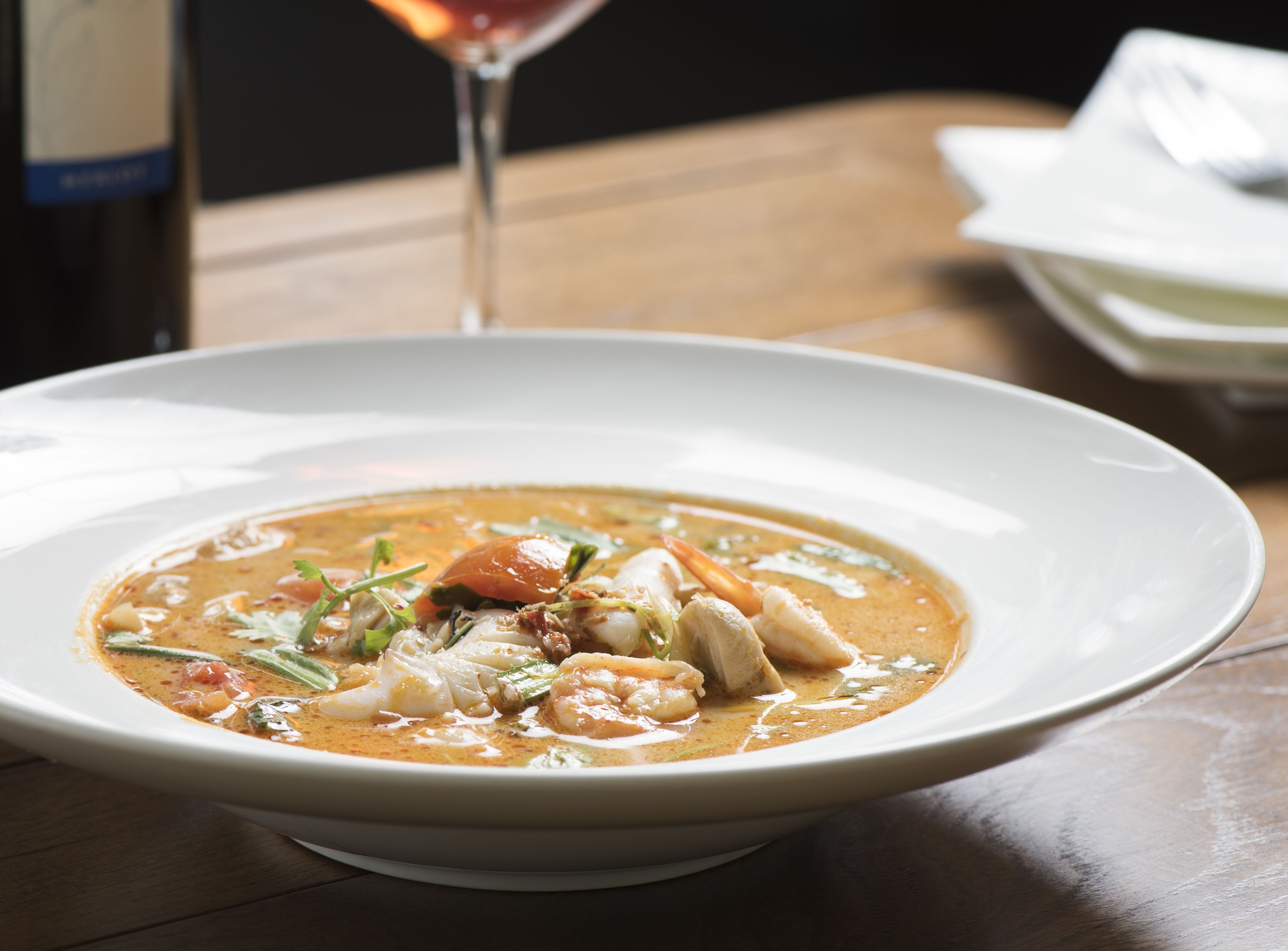 Un tazón de sopa de mariscos sobre una mesa de madera, con una copa de vino y una servilleta al fondo.