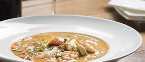 Un tazón de sopa de mariscos sobre una mesa de madera, con una copa de vino y una servilleta al fondo.