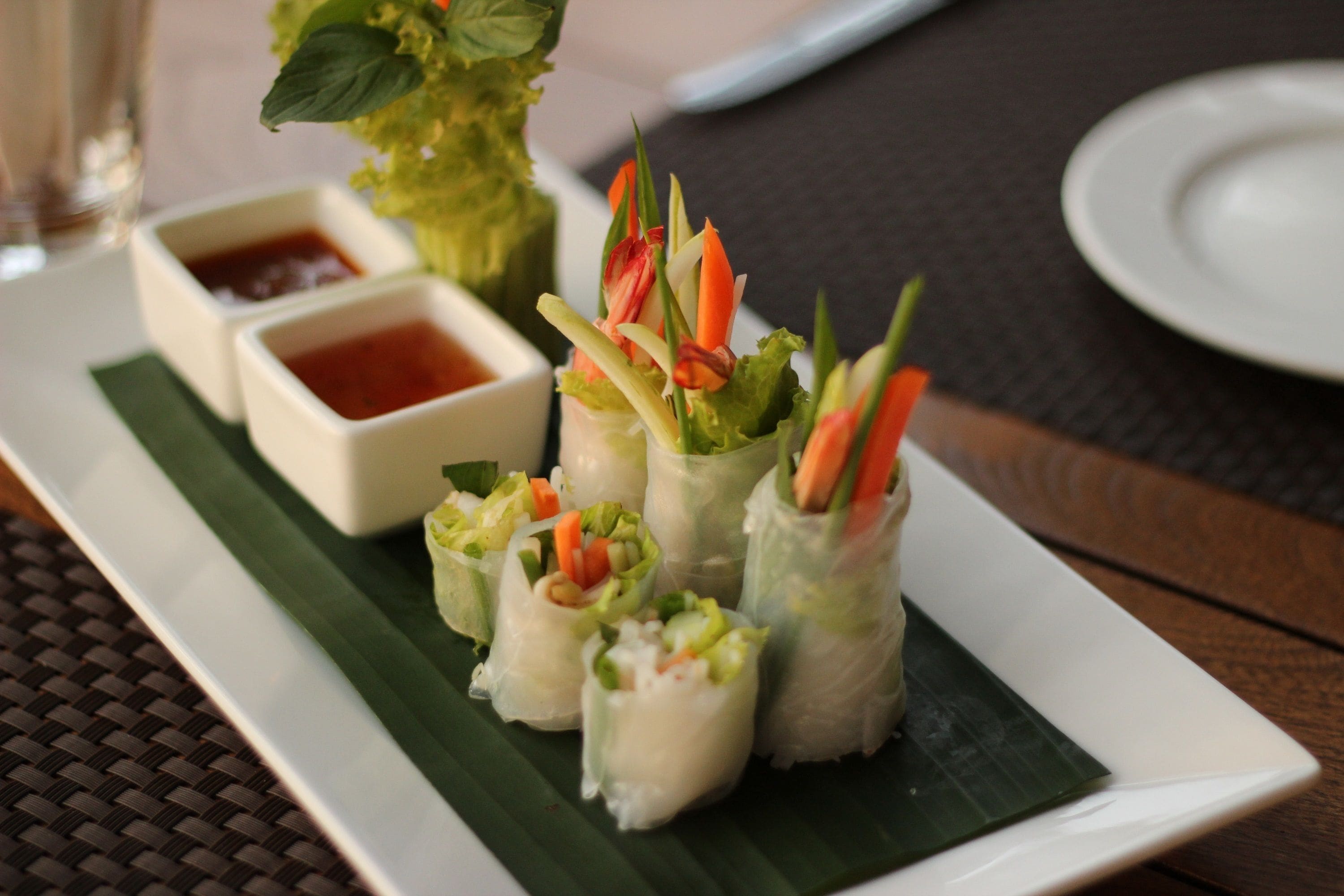 Un plato de rollitos vietnamitas con salsas para mojar, servido sobre un plato blanco con una hoja verde debajo.