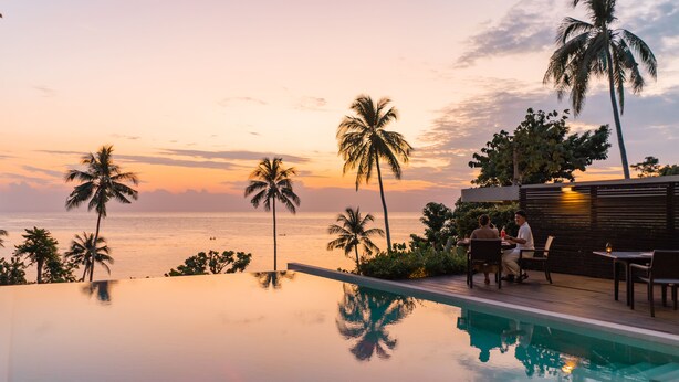 Une piscine à débordement offrant une vue sur l’océan et les palmiers au coucher du soleil.