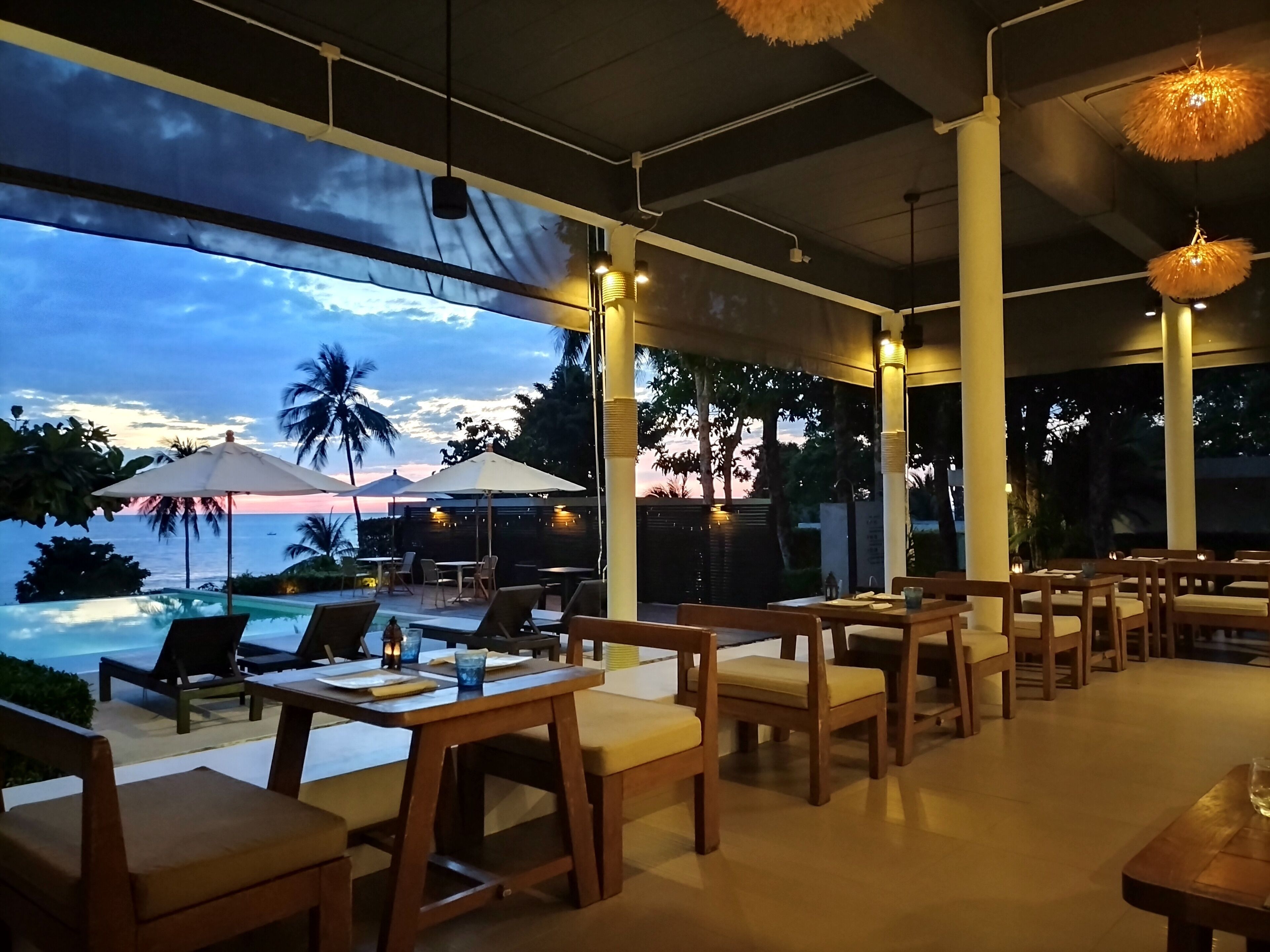 Un restaurante con terraza al aire libre que da a una piscina y vistas al océano, con mesas y sillas de madera y grandes sombrillas.