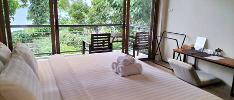 A hotel room with a large bed, a desk with a laptop, and a view of greenery outside.